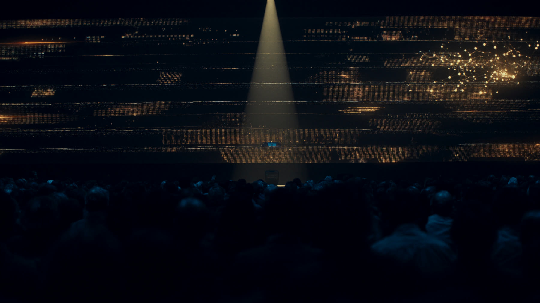Auditorio a oscuras frente a un escenario iluminado por un haz de luz y pantallas con datos en tonos dorados